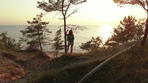 Young Woman Enjoys Beautiful Scenic Sunset at Baltic Sea Standing at Tree on Hill. Slow Aerial Orbit