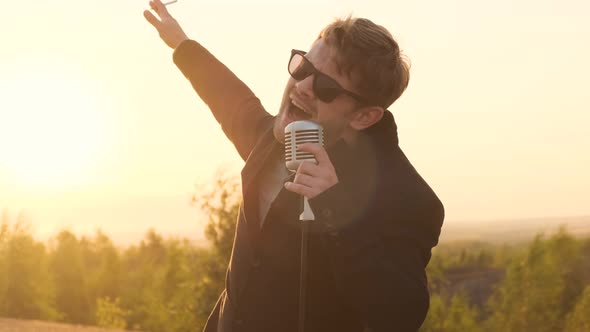 Man in Black Glasses and Black Jacket Sings Song Into Shiny Vintage Retro Microphone in Nature