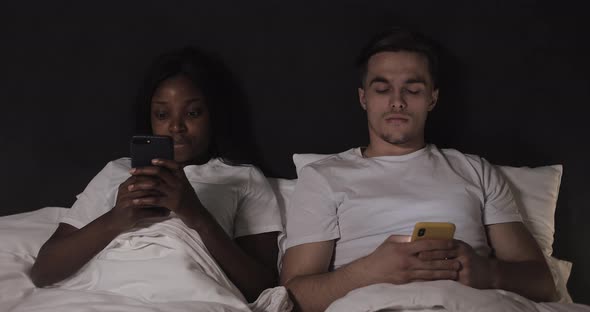 Multi-ethnic Couple Using Smartphones Lying in the Bed at Night. Man Peeks on His Woman s Phone, She