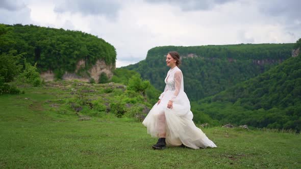 A Girl in a Wedding Dress and Black Boots Goes on the Lawn in the Mountains