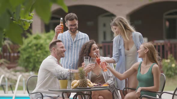 Group of happy young people cheering with cider by the pool in the garden