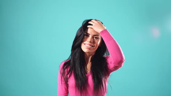 Smiling Brunette Woman Playing with Her Hair and Looking at the Camera Over Turquoise Background.