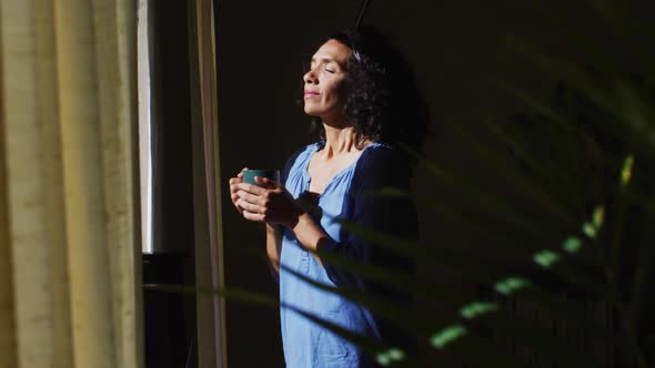 Mixed race woman holding coffee cup while standing near the window at home