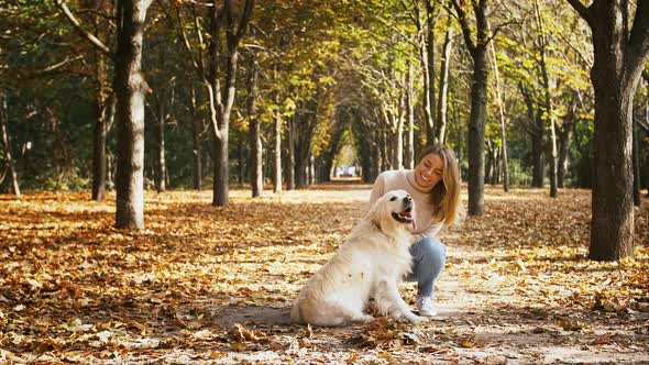 Young Lady in Casual Outfit is Squatting on Alley of Autumn Park Smiling Stroking Her Dog Labrador