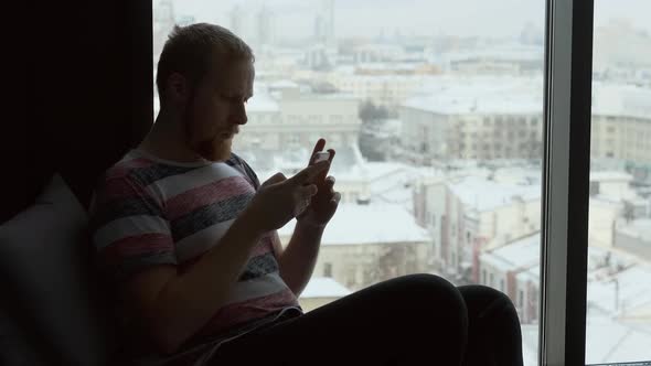 a man with a smartphone sitting by the window overlooking the city