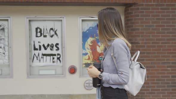 Woman walks parallel to building with black lives matter poster, looks at poster, side view, wearing