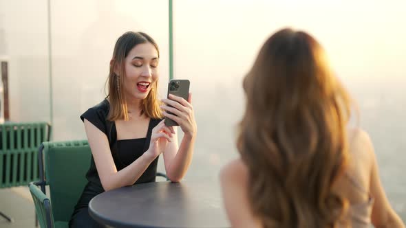 4K Asian woman friends using smartphone photographing together at rooftop restaurant at sunset.