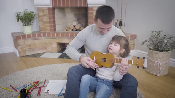 Portrait of Young Caucasian Handsome Man Teaching Daughter To Play Ukulele. Pretty Little Girl