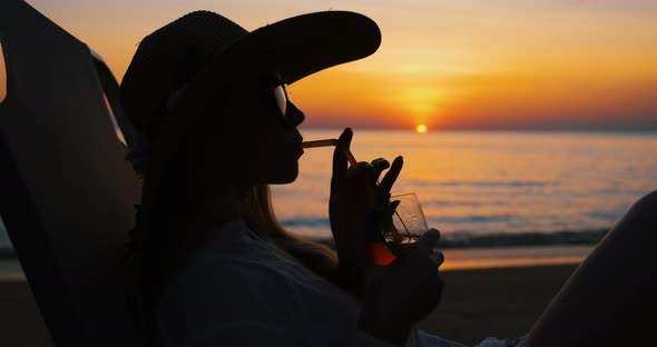 Silhouette of Woman Drinking Cocktail on the Beach at Sunset Lying on