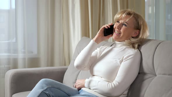 Blondehaired Woman Talks Via Smartphone and Smiles on Sofa