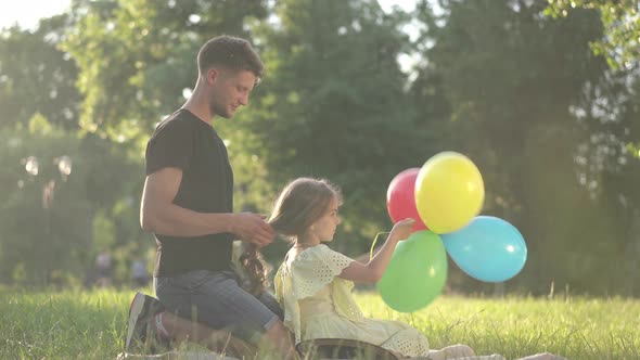 Side View Relaxed Daughter Playing with Colorful Balloons As Young Father Braiding Hair Sitting on