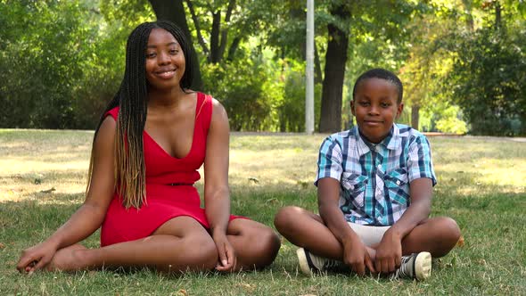 A Young Black Mother and Her Son Sit on Grass in a Park and Smile at the Camera