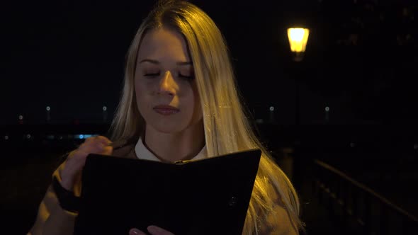 A Young Beautiful Woman Writes Into a Notebook in an Urban Area at Night