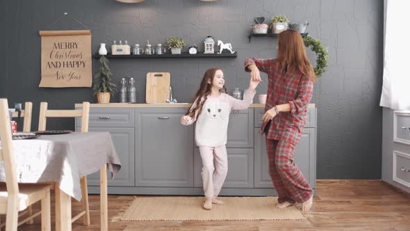 Joyful Young Beautiful Woman with a Little Girl Dancing in the Kitchen Wearing Pajamas in the