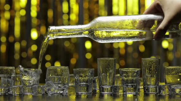 Barman Pour Frozen Vodka From Bottle Into Shot Glass. Ice Cubes Against Shiny Gold Party Background
