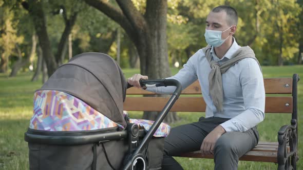Young Caucasian Man in Covid Face Mask Rocking Baby Carriage in Summer Park. Portrait of Handsome