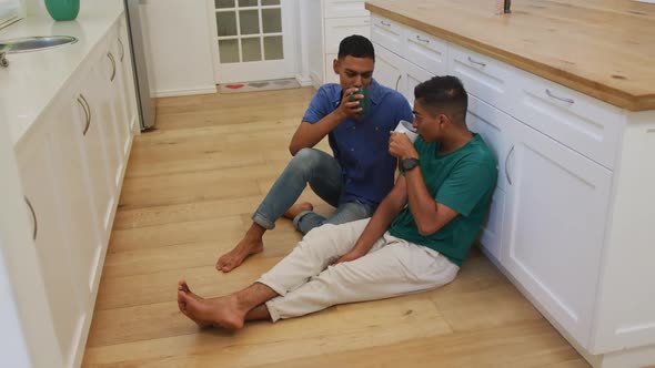 Happy mixed race gay male couple sitting on kitchen floor talking and drinking coffee