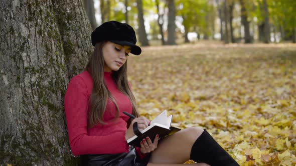 Young Girl Leaned on an Autumn Tree Writing Down Thoughts in a Notebook