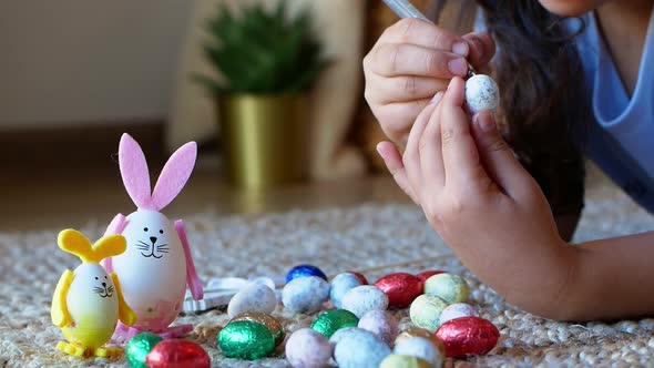A girl draws a pattern on an Easter egg next to an egg in the shape of a rabbit alt