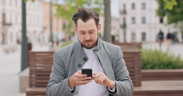 Young Man with Mustaches and a Beard Sitting on a Bench in the Square Scrolling the Smartphone