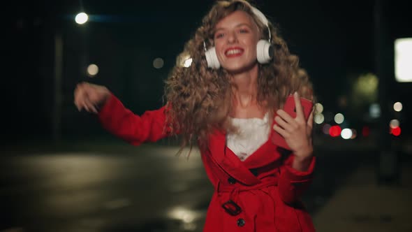 A Smiling Curly Woman in Headphones is Listening Music and Funny Dancing in City Street