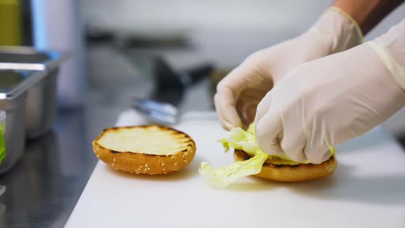 Hands in latex gloves put lettuce on top of fried bun. 