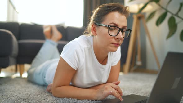 Woman with Glasses is Lying on the Floor and Working on a Laptop