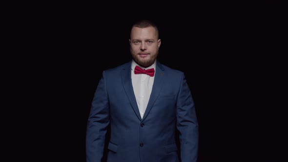 An elegant man in a blue tuxedo and a red tie smiles sweetly against a black background