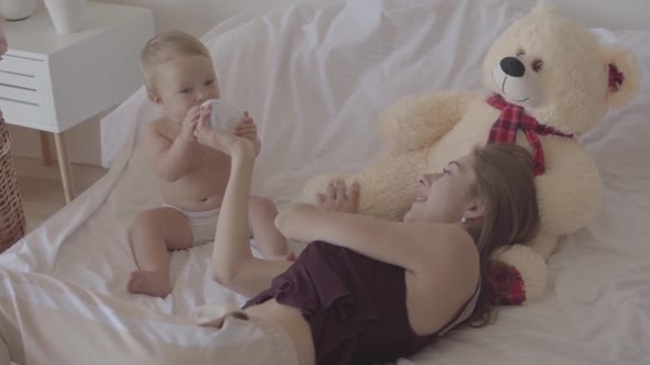 Young Woman Sitting with Her Baby Girl Who Playing with Baby Bottle Sitting in the Bed, Bid Toy Bear