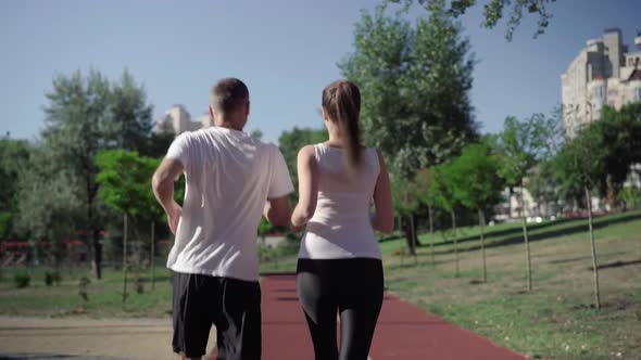 Back View of Adult Couple of Joggers Running Along Track on Summer Morning. Camera Follows Fit