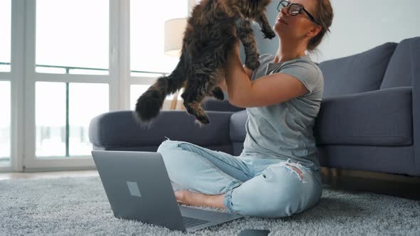 Casually Dressed Woman Sits on a Carpet with a Laptop Holds on Her Knees and Strokes a Fluffy Cat