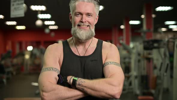 Smiling gray-haired respectable man with a beard and tattoos posing in the gym