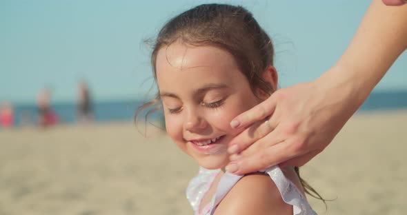 Mother Puts Sunscreen on a Little Girl's Face While Sitting on the Beach