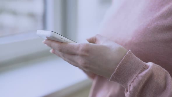 Pregnant Woman Standing by Window With Smartphone in Hands and Scrolling Screen