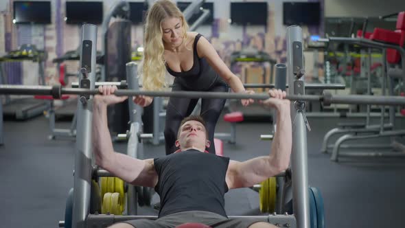 Fit Slim Blond Personal Trainer Helping Sportsman Lifting Barbell in Gym Lying on Exercise Bench