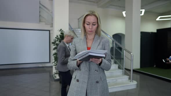 Thoughtful Concentrated Businesswoman Walking Tapping Fingers on Paperwork with Colleagues Passing