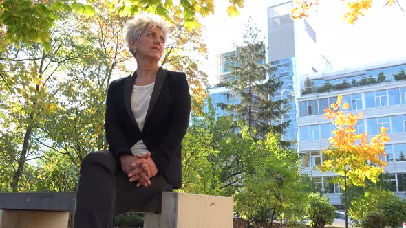 A Middle-aged Businesswoman Sits on a Bench in a Park and Looks Around - an Office Building