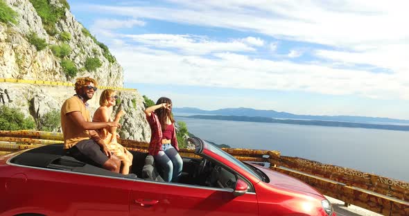 Multi ethnic friends partying in red convertible parked on side of coastal road