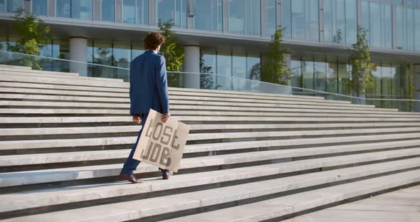 Young Man in Suit Holding Lost Job Cardboard Sign Walking Outside Business Center