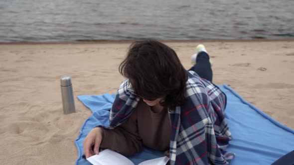 Caucasian Woman with Glasses Reads a Book Lying on a Sandy Beach