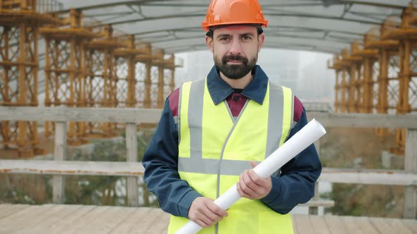 Portrait of Male Builder Wearing Helmet and Vest Standing Outside Construction Site Holding