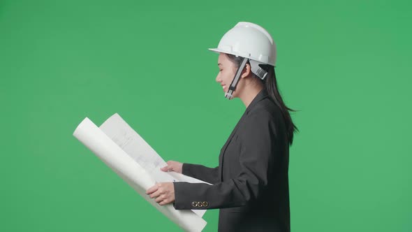 Side View Of Smiling Asian Female Engineer Looking At Blueprint While Walking On Green Screen