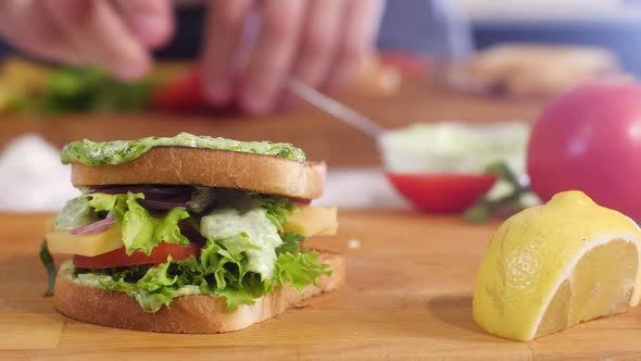 man puts pesto sauce on bread, ripe tomato slices and onion slices, and cheese.