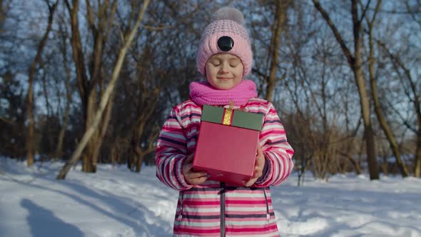 Smiling Child Kid in Snowy Winter Forest Park Looking at Camera Holding Christmas Present Gift Box