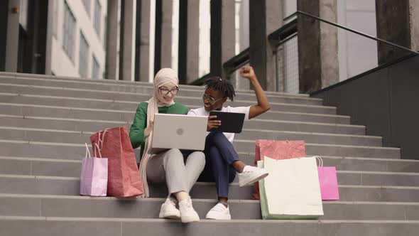 Multiethnical Girls Sitting on Steps of a Modern Building Rejoicing with Online Orderafter Shopping