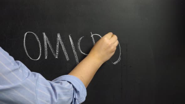 A woman's hand writes the word Omicron in white chalk on a black board.