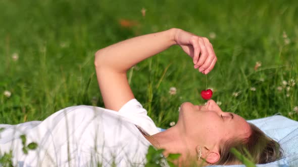 A Young Girl Lies on the Green Grass and Tastes a Ripe Red Cherry