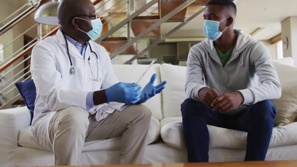 African american senior male doctor giving covid test to male patient in home, wearing face masks