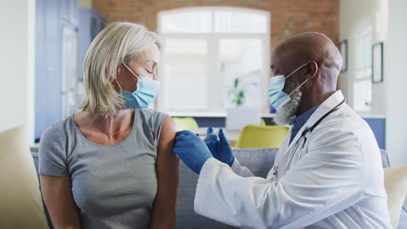 Happy senior diverse woman and doctor wearing face masks in living room sitting on sofa, vaccinating