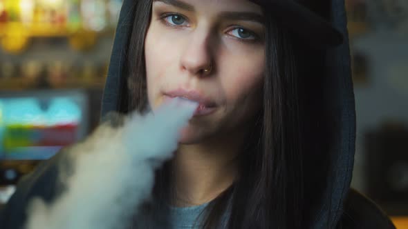 Young Pretty Woman in Cap Smoke an Electronic Cigarette at the Vape Shop. Hip-hop Style. Closeup.